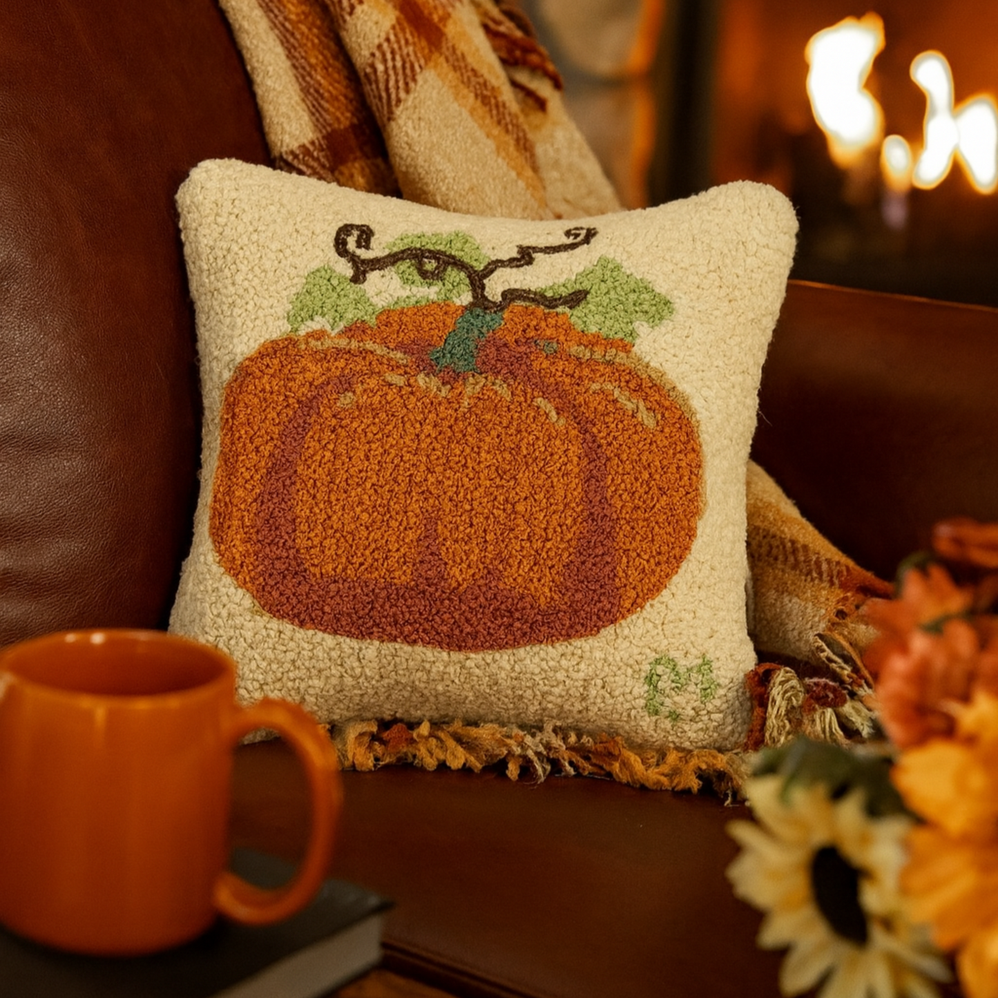 Decorative pillow with a pumpkin design on a couch next to a mug and books, with a fireplace in the background.