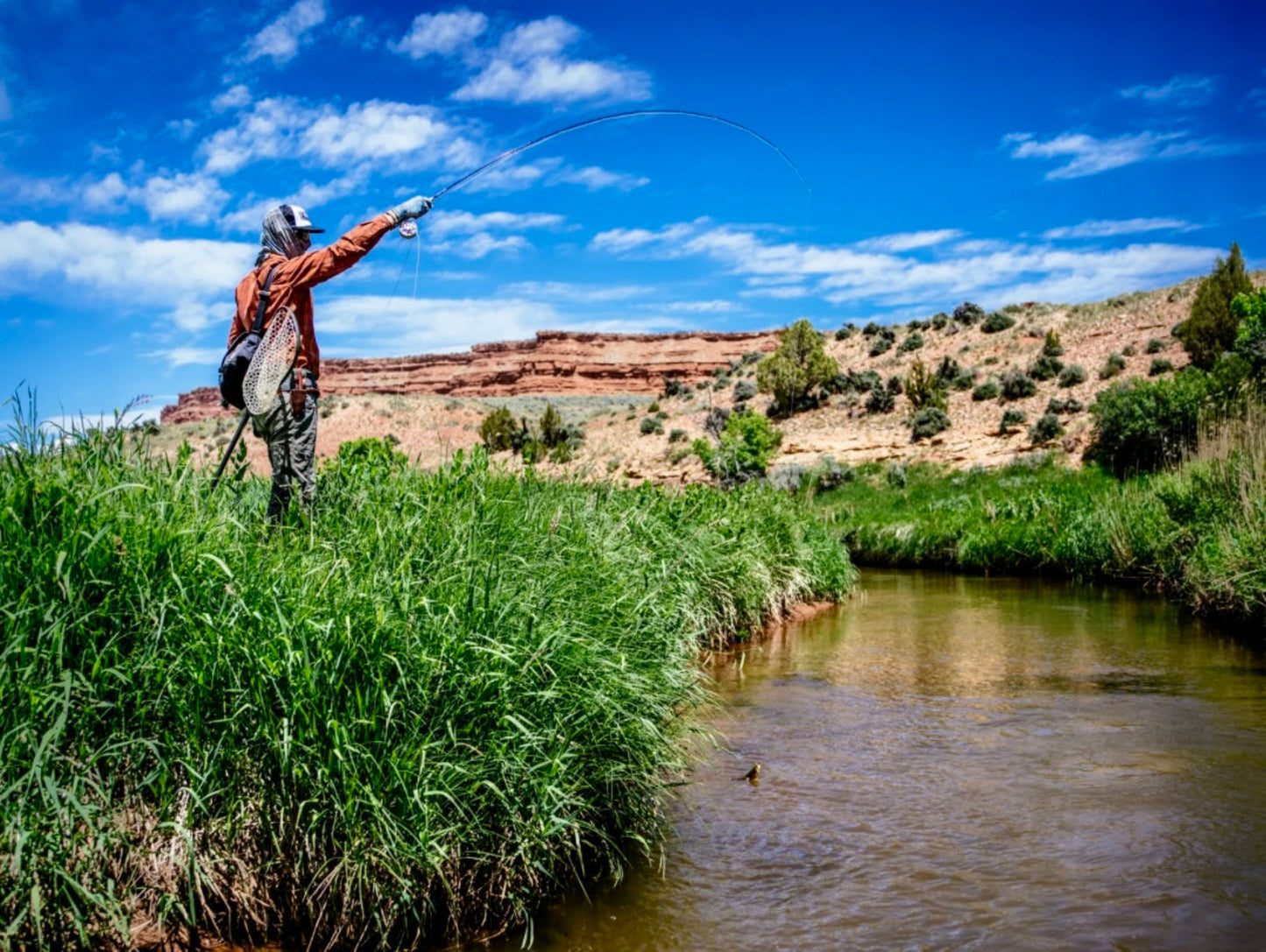 Person fly fishing in a river with a scenic landscape and blue sky.